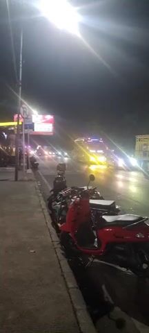 The video shows a nighttime street scene. In the foreground, several motorcycles are parked along a sidewalk. One red scooter is prominent. In the background, a bus with flashing orange lights is driving down the road, which is illuminated by streetlights and the headlights of passing vehicles. The atmosphere is dark and the road appears wet, possibly from rain, reflecting the lights. A sign with a red circle and a diagonal line is visible on a pole near the sidewalk.