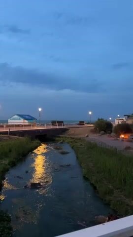 The video shows a serene view of a small river flowing towards the ocean under a twilight sky. The river is bordered by green grass and rocks. A bridge crosses the river, with a building featuring a blue roof situated beyond it. Streetlights illuminate the area along the promenade, which leads to the beach. The scene is calm and peaceful, with no people visible. The water reflects the yellow glow of the streetlights, creating a tranquil ambiance.