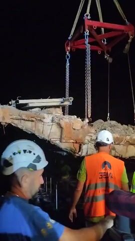 The video shows a nighttime scene with a large, broken concrete structure being lifted by a crane. Two men in hard hats and safety vests are visible in the foreground, looking towards the lifted debris. One man wears a blue shirt and a white hard hat. The other man, facing away from the camera, wears an orange safety vest with a logo that appears to be "GEN" and a white hard hat. The debris itself is large and appears to be a section of a bridge or overpass, with exposed rebar and broken concrete. The background is dark, indicating nighttime, with some lights visible in the distance, possibly from other construction equipment or a tunnel entrance. The crane's lifting apparatus, with chains and a red frame, is prominent at the top of the frame. The overall impression is of a demolition or cleanup operation after a structural failure.