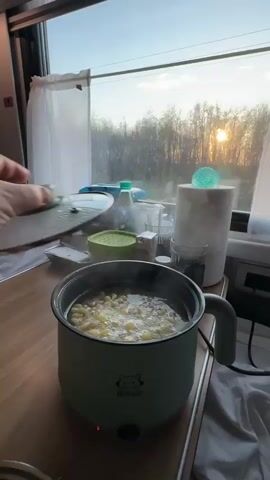 The video shows a person cooking in what appears to be a train compartment. A small, light green electric pot sits on a wooden table with a steamer insert placed on top. The pot contains boiling water with something inside, possibly pasta or small vegetables. The steamer insert holds cooked, yellowish dumplings and sliced sausage. A hand places the lid on the steamer insert.  The background includes a window offering a view of a sunrise over a wooded area. Other items on the table include a pillow, a box of wet wipes, a bottle of hand sanitizer, glasses, a bowl of green powder, and a roll of paper towels. The bed is rumpled in the background.