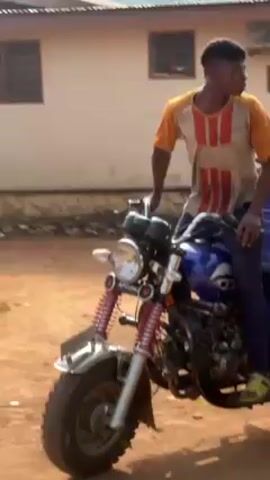 A young man is seated on a blue three-wheeled motorcycle with "APSONIC" printed on the cargo bed. The bed is loaded with tree trunks and branches. The motorcycle moves forward on a dirt road. The background shows buildings and fences.