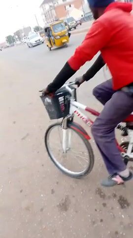The video shows a man in a red shirt and blue beanie riding a bicycle on a paved road next to a drainage ditch. There is a basket on the front of the bicycle. The man is traveling in the same direction as the camera. Another man walks next to the drainage ditch, holding a bag or container. He looks at the camera at one point. Several vehicles, including cars and a tricycle, are visible on the road in the background, as well as buildings and power lines. The sky is overcast. The ground appears to be concrete or asphalt. The video ends with a view of the road ahead.