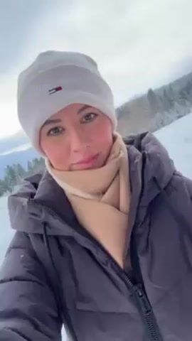A woman wearing a white Tommy Hilfiger beanie, beige scarf, and a dark grey puffer jacket stands in a snowy landscape. Trees are visible in the background. The sky is overcast. The camera angle is from a selfie perspective. The woman is looking directly at the camera with a slight smile. The overall tone is calm and serene.
