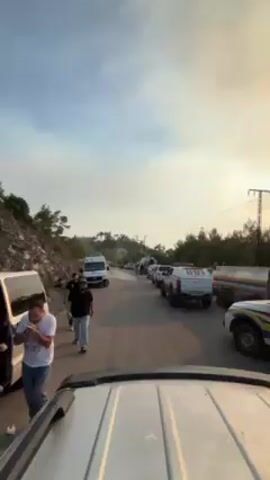 Here is a forensic description of the video:

The video, taken from inside the windshield of a light-colored vehicle, shows a traffic jam of various vehicles on a narrow, paved road bordered by a rocky hillside on the left and trees on the right. Several people stand around or walk alongside the vehicles. The mix of vehicles includes standard cars, vans, and specialized trucks, some of which appear to be tankers or work trucks, potentially involved in emergency services due to their size and utility. The sky above is a light blue color, tinged with a hazy, golden hue, suggesting either early morning or late afternoon. This haze is particularly noticeable in the distance, which might be indicative of smoke or other airborne particles. The terrain on either side of the road is varied with foliage, and a power pole is visible along the side of the road. The overall impression is one of a busy, possibly critical or emergency-related situation in a rural or semi-rural area.