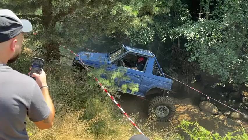 A person wearing a white baseball cap and a gray t-shirt films an off-road vehicle with their phone. The vehicle, a modified blue SUV with large tires, is navigating a steep, muddy incline surrounded by trees and foliage. Red and white caution tape marks the path. The vehicle's tires are kicking up a significant amount of dirt and dust as it attempts to climb the obstacle. Another person, wearing a black t-shirt with white lettering, is partially visible to the left of the frame, also observing the event. The overall environment appears to be a wooded area during daytime.
