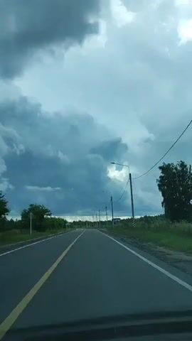 A first-person perspective video shows a paved road with a yellow line down the middle, stretching into the distance. The road is lined with green trees and bushes on either side. The sky is filled with dark, dramatic clouds, suggesting an impending storm. Several lampposts are visible along the right side of the road. The scene appears to be a rural or semi-rural area.