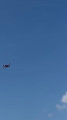 A small, red and white airplane is captured in mid-flight against a clear blue sky. The airplane appears to be performing aerobatics, as evidenced by a white smoke trail that extends from its tail, forming a curved shape in the sky. The sun is high, suggesting it is daytime, and there are a few wispy clouds visible near the bottom of the frame, indicating fair weather. The scene is set in an open area, likely rural, given the vast expanse of sky and lack of prominent structures. The image conveys a sense of freedom and excitement associated with flight and aerial displays. No visible text is present in the media file.