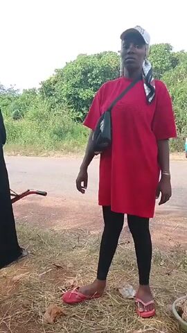A woman in a red shirt, black leggings, and red flip-flops is standing on a patch of dry grass, adjusting her cap and talking.  She has a black bag slung across her body.  A person in a black abaya and headscarf stands to her left. A red object, possibly a bicycle part, lies on the ground nearby.  A paved road stretches in front of them, and a background of green trees and foliage completes the scene.