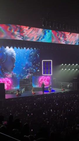 The video shows a concert performance. A performer stands on stage center, facing the audience. Several backup dancers are positioned around the stage. Large screens display vibrant, abstract visuals and an image of a person with headphones. The stage is lit with bright lights, and the audience is visible in the foreground, many holding up their phones.