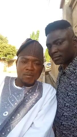 Two men are standing close together outdoors, taking a selfie-style video. The man on the left wears a white long-sleeved shirt with a grey and white patterned vest and a brown hat. The man on the right wears a black and white patterned button-up shirt. The man on the left raises two fingers in a peace sign gesture. They both look at the camera. Behind them is a wall, some trees, and a parked car in the distance. The sky is overcast.