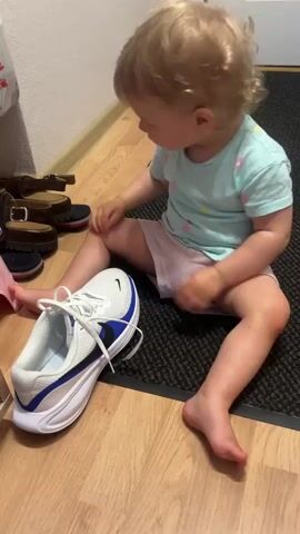 A young child with blonde curly hair, wearing a light blue t-shirt with small colorful dots and a pink diaper or pull-up, sits on a dark gray doormat in front of a shoe rack. The child is looking down at a white and blue athletic shoe with white laces. The child's left foot is extended towards the shoe, and their right leg is bent. To the left of the child, on the shoe rack, are several pairs of shoes, including brown sandals and dark blue shoes. A pink object is partially visible to the left of the child. The floor is made of light brown wood. The lighting suggests an indoor environment.