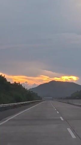 The video shows a highway with a sunset in the background. The sky is cloudy, with orange and yellow hues from the setting sun visible between the clouds and behind a mountain. The highway is empty and appears to be a multi-lane road with white lane markings. The sides of the highway are lined with greenery. The camera is moving forward, suggesting it is from a vehicle traveling on the highway.