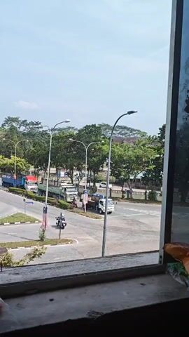 The video captures a view from a window overlooking a street intersection. Several large trucks are visible, some on the road and some stopped. A motorcycle is also present at the intersection. Trees line the street, and a clear blue sky is overhead. The foreground shows the windowsill, which appears to be made of concrete and has some debris on it. The overall scene suggests a somewhat industrial or commercial area.