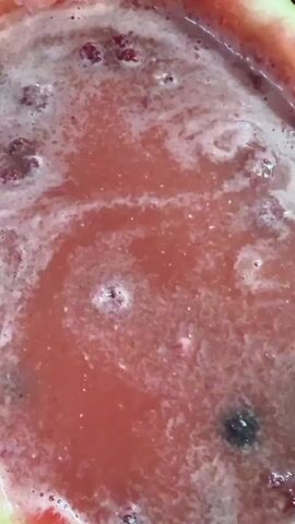 A close-up shot shows the inside of a hollowed-out watermelon. The flesh of the watermelon has been scooped out, creating a bowl filled with pink juice and some small dark seeds or fruit pieces. The surface of the juice is foamy and swirling. In the center of the frame, a metal spoon is used to stir the liquid and some of the fruit pieces. The watermelon rind is visible around the edges of the frame, showing its green and pink exterior. The lighting is dim, and the focus is on the contents of the watermelon.