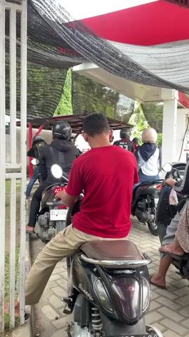 The video shows a line of motorcyclists at what appears to be a gas station. The camera is positioned behind a person wearing a red shirt and khaki pants, who is sitting on a black scooter in the foreground. The person's face is not visible. The scooter has a brown seat and a Honda logo. Behind this person, other motorcyclists are waiting in line. The people in the line are wearing helmets and casual clothing. The environment is an outdoor area with a covered structure overhead, possibly a canopy or roof of the gas station. The canopy is partially covered with black netting. There are trees and foliage visible in the background. The ground is paved with interlocking bricks. The scene suggests a normal day at a busy gas station.