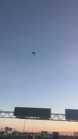 The video shows a plane flying overhead against a blue sky with a hint of orange at the horizon, presumably during sunrise or sunset. The view is from a moving vehicle on a highway, with multiple lanes visible. There are cars driving on the highway and overhead signage structures. Streetlights are spaced along the side of the road.