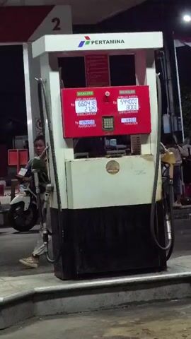 The video shows a Pertamina gas pump at night. The pump has two displays showing fuel prices and quantities. On the left, "DEXLITE" is priced at Rp 24,200 per liter, with 27.29 liters dispensed for a total of Rp 660,418. On the right, "BIO SOLAR" is priced at Rp 6,800 per liter, with 80.00 liters dispensed for a total of Rp 544,000. A person in a black shirt and grey pants stands to the left of the pump, and a motorcycle is visible behind them. Another person is partially visible to the right of the pump. The scene appears to be a gas station with overhead lighting.