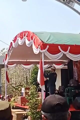 A man wearing a dark jacket, dark pants, and a light-colored hat stands on a stage and holds an Indonesian flag. He appears to be speaking into a microphone. The stage is covered by a tent with red and white trim. In the background, there are trees and other people. The scene is outdoors and appears to be a daytime event.