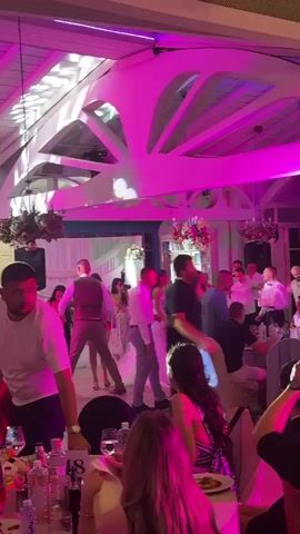 A wedding reception is underway with guests dancing and dining. The room is lit with vibrant pink and green lights, illuminating the white ceiling beams and hanging floral arrangements. Several people are on the dance floor, including a bride in a white wedding dress and other guests in formal attire. Some guests are seated at tables, enjoying their meals. The overall atmosphere appears festive and celebratory.