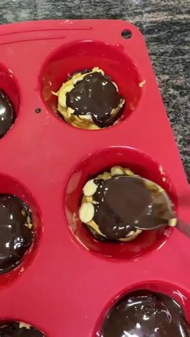 A close-up shot shows a red silicone mold with six round cavities, each containing a pastry. A spoon is in the process of drizzling melted dark chocolate over one of the pastries. The pastries appear to be cream puffs, with a light-colored filling peeking out from between layers of dough, and they are already partially covered in chocolate. The mold is placed on a countertop with a speckled pattern. The lighting is bright, and the focus is sharp.