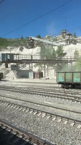 The video shows a train passing by an industrial site, likely a quarry. The train cars are green and brown, with visible markings like "65070963" and "62394225." The quarry features a large white hillside with various industrial structures including conveyor belts and crushing equipment. Dust is visible in the air, likely from the quarry operations. The sky is clear and blue. The viewpoint is from within another train, looking out the window.