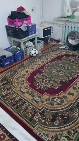The video shows a cluttered room with a large, ornate rug dominating the foreground. To the left, several plastic crates are stacked on a table, filled with various items, including clothes. A soccer ball rests on the floor near the crates. In the background, a microwave oven sits on a counter next to a radiator. A black fan is positioned to the right of the radiator. The room appears to be in a state of disarray, with items scattered around. The lighting is dim, and the overall atmosphere is somewhat cramped.