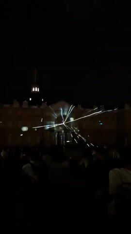 A wide shot shows a building at night with a light show projected onto its facade. White lines of light emanate from a central point on the building, creating a starburst or radiating pattern. Smaller dots of light, some appearing green, are scattered across the building. In the foreground, a crowd of people is visible, mostly silhouetted against the light show, suggesting they are facing the building and watching the display. The sky is dark, indicating it is nighttime.