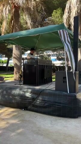 The video shows a DJ under a canopy playing music for a crowd gathered outdoors. The DJ, wearing sunglasses and headphones, stands behind a black booth with speakers. A white and green striped flag hangs from the canopy. The crowd is diverse, with people of different ages and clothing styles dancing and socializing under a shaded area with palm trees in the background. The ground is sandy or gravelly. The sky is clear and blue.