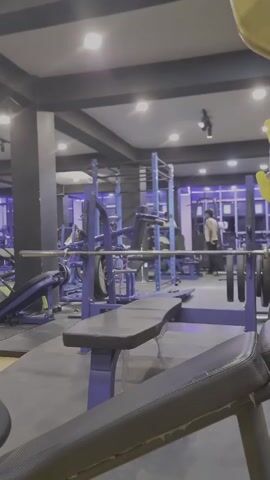 Here is a concise, forensic description of the scene:

The video shows the interior of a gym. In the foreground, there's a bench press setup with a padded bench, a barbell, and several weight plates on each side. The bench and its frame are predominantly blue.  The floor around the bench is covered with black mats. Other gym equipment, also mostly blue, is visible in the background.  A person wearing a light-colored tank top is walking in the background. The gym is lit with several ceiling lights, and the overall color scheme is a mix of gray, black, and blue.