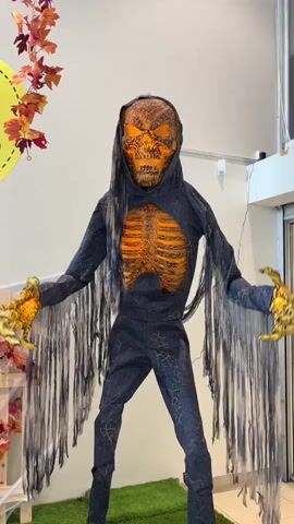 A tall, Grim Reaper-like Halloween animatronic stands in the center of the frame. It has a hooded, dark blue costume with a cracked pattern. The chest is exposed, revealing orange rib bones. The mask is a grinning skull with glowing orange eyes. The animatronic has long, tattered black sleeves and large, gold, claw-like hands. It stands on a green artificial turf platform. To the left, there are wooden crates decorated with artificial autumn leaves and black plastic spiders. A yellow circular sign with red leaves and the text "Spar 20%" is visible. The background consists of a plain wall and a glass storefront. The video briefly pans and zooms out, showing more of the surrounding store environment.