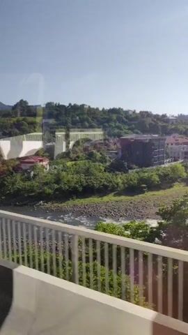 The video shows a view from a moving vehicle, likely a car or bus, traveling over a bridge. The bridge has a white and gray barrier in the foreground. Below the bridge is a rocky river with flowing water. Across the river, there is a lush green hillside covered in trees and vegetation. Several buildings are visible on the hillside, including houses, a multi-story apartment building, and what appears to be a large white structure with multiple sections that could be a dam or a complex of buildings. The sky is clear and blue. The overall impression is a scenic view of a town or city nestled in a valley beside a river and surrounded by hills.