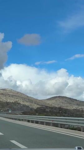 The video shows a view from a car driving on a highway. On the side of the highway is a mountainous terrain with scattered vegetation. The sky is blue with puffy white clouds. The car is moving, as evidenced by the changing scenery. A guardrail runs along the side of the highway. There is a road sign visible. The overall scene depicts a drive through a mountainous region on a partly cloudy day.