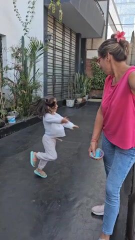 The video shows a young girl and an older woman in an outdoor space. The girl, wearing a white long-sleeved shirt, light gray pants, and colorful sandals, is dancing and running. She has dark hair and a bright smile. The woman, wearing a pink sleeveless shirt, blue jeans, and gray sneakers, is holding a small container and watching the girl. The woman has a flower in her hair. The background includes a building with a black-framed window, various potted plants, and some greenery. The ground is a dark, paved surface.