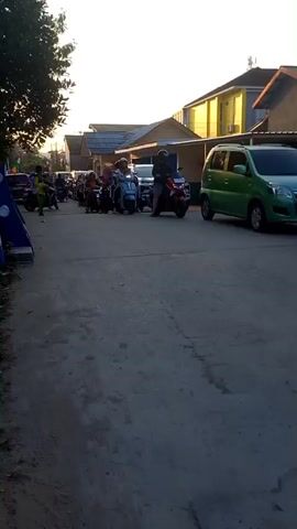 A street scene with numerous motorcycles and a few cars. The sun is setting, casting long shadows. People are riding on the motorcycles, some with passengers, including children. Buildings line the street, and trees and palm trees are visible. A flag with red and white stripes is flying near a small building. A sign with text in a foreign language is visible on the back of a car. The overall atmosphere suggests a busy evening, possibly during a local event or commute.