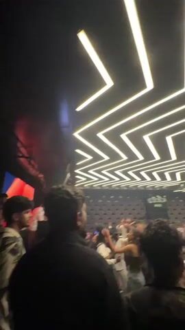 A video showing a crowd of people in a dark room with bright, yellow zigzag lights on the ceiling. There are men and women dancing in the room, some people are wearing white shirts, some people are wearing patterned shirts, and a woman is wearing a dark top with blue jeans. In the background, there is a large screen with a red and blue flag visible. There is a man wearing a black shirt with his back to the camera, standing and looking at the crowd.