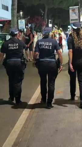 Three individuals wearing police uniforms with "POLIZIA LOCALE" visible on their backs are walking down a street at night. They are walking away from the camera, towards a crowd of people. The street appears to be a pedestrian area with shops and trees lining the sides. The crowd consists of various people, including adults and possibly children, some of whom are looking in the direction of the police officers. The lighting suggests it is evening or night, with streetlights and lights from the surrounding businesses illuminating the scene. The ground is paved, and there are some fallen leaves on the pavement. The police officers are wearing dark uniforms, caps, and appear to have utility belts.