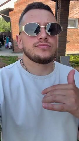 This is a close-up, selfie-style video of a man with short brown hair, a beard, and sunglasses with gold frames. He is wearing a light-colored t-shirt and a silver chain necklace. He is looking directly at the camera with a neutral expression and a slight smirk. His right hand is raised, with his thumb extended upwards and his fingers pointing towards the camera.

The background suggests an outdoor setting, likely a backyard. There is a brick building with a roof structure behind him. A car is visible to the left, and some chairs and gardening equipment are scattered around. The lighting indicates it is daytime, possibly late afternoon, with natural sunlight present.

The video appears to be a short, casual clip, possibly a greeting or a social media update. The man's pose and expression convey confidence and a relaxed demeanor. The setting seems to be a residential area in Tulinskiy, Russia, given the provided location context. No visible text is present in the media.
