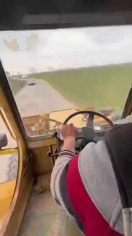 From the perspective of inside a yellow, old piece of heavy machinery, likely a bulldozer, a man is driving on a narrow paved road through a rural area. He is wearing a grey, red, and white striped sweatshirt and is holding the steering wheel with his right hand.  Through the windshield, another vehicle is approaching in the distance.  To the right of the road is a green field, and some buildings are visible in the background. The weather appears overcast.