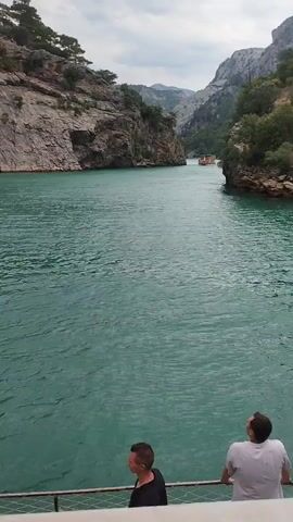 The video shows a boat tour on a narrow turquoise body of water surrounded by steep, rocky cliffs covered in sparse vegetation. The boat appears to be moving forward, as indicated by the water churning at the stern. On the deck of the boat, several passengers are visible, some standing and looking out at the scenery, while others are seated. The passengers include adults and children, and they are dressed in casual summer clothing. Some luggage, including backpacks and bags, is also present on the deck. The sky is overcast with some patches of blue visible. In the distance, another boat can be seen traveling in the same direction. The overall impression is one of a scenic boat trip through a canyon.