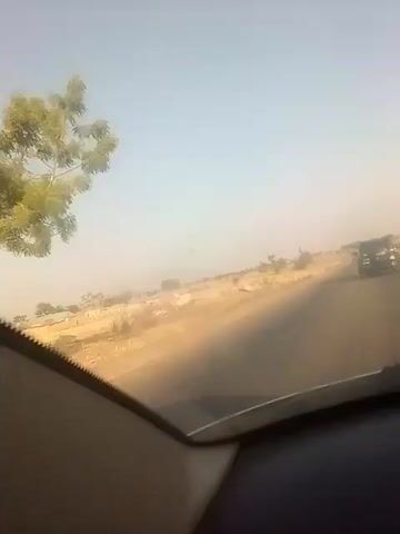 The video shows a view from inside a moving car looking out over a two-lane road through a dry, dusty landscape under a bright, hazy sky. There are some small trees and scrub brush along the side of the road, and what appear to be some buildings in the distance. A car and a motorcycle are visible on the road ahead. The interior of the car is visible in the foreground. Music is playing.
