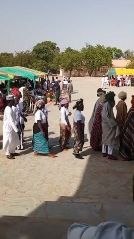 The video shows a procession of people, primarily men in traditional attire, walking in a courtyard. The men are wearing turbans and long, flowing robes with varying patterns and colors. Some are carrying umbrellas, including a large red one held over a central group. The procession appears to be moving from right to left in the frame. In the background, there are tents set up, with other people gathered, some seated and some standing. Trees and buildings are visible beyond the tents, suggesting an outdoor event in a settled area. The ground is paved with light-colored material, possibly concrete or packed earth. The overall lighting indicates it is daytime, likely under a bright sun. Several individuals are visible in the foreground, facing away from the camera, also appearing to be spectators or participants.