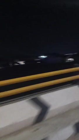 A nighttime view from a vehicle moving across a bridge. The sky is dark, with a few faint lights visible in the distance. The bridge has a concrete barrier with yellow and white guardrails. Streetlights illuminate the road, and traffic signals are visible, showing green lights. The camera captures the movement of other vehicles on the road.