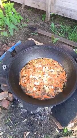 A top-down shot focuses on a large black cast iron pot, likely a kazan, set over an open fire. The pot is filled with food that appears to be pilaf. Visible ingredients include julienned carrots, meat, and several whole cloves of garlic scattered on top. The food is bubbling and cooking in oil. The pot is resting on a makeshift stove constructed from rough stones and concrete blocks. The surrounding environment is outdoors, with dirt, dry leaves, and some green grass visible. Wooden logs and a blue tool are also present in the background. The lighting suggests it is daytime.