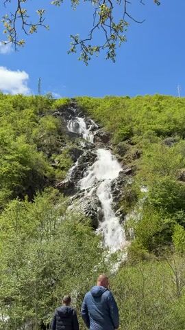 The video shows a tall waterfall cascading down a verdant, tree-covered hillside under a clear blue sky with a few wispy clouds. Two men stand at the bottom of the view, looking up at the waterfall. One is wearing a blue jacket, and the other a dark jacket. In the upper corners of the frame, tree branches with budding leaves are visible, slightly obscuring the sky. Electrical towers are visible on the top of the hill near the waterfall.