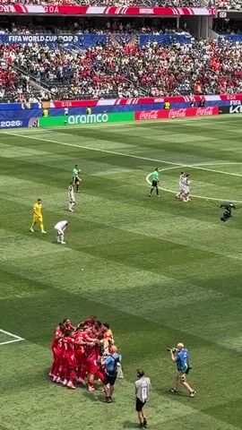 The video shows a soccer game from a high vantage point. In the foreground, a group of players in red uniforms are celebrating, huddled together in the center of the field. Several photographers and camera operators are positioned around the group, some of whom are taking pictures and recording video. In the middle ground, a few players in white and yellow uniforms, along with referees in green and black uniforms, are scattered across the field, appearing to have concluded their activity. In the background, a large stadium filled with spectators wearing red and white is visible, with various advertisements displayed along the stadium walls. The field itself is a vibrant green with distinct white lines.