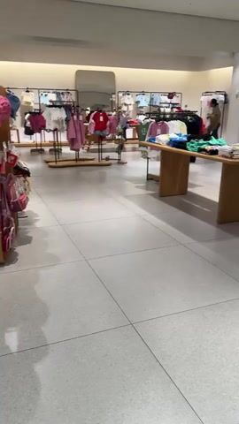 The video shows the interior of a clothing store. In the foreground, a white short-sleeved shirt with a tag is displayed on a stand. To the right, other clothing items are visible, including green t-shirts and black shorts. In the background, a young child wearing a camouflage t-shirt and dark pants stands facing away from the camera, looking at clothing racks. Further back, a woman with dark hair is seen standing near a clothing rack. The store appears to be well-lit with white walls and a tiled floor. Multiple clothing racks displaying various garments are arranged throughout the space. Some of the clothes appear to be children's wear. A large mirror is visible on the far wall.