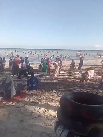 The video shows a crowded beach on a sunny day.  People are wading in the shallow water, swimming, and walking along the shore. Some people are wearing swimming rings.  Several groups of people are walking on the beach in the foreground.  Seaweed is scattered along the sand.  A man sits on the sand in the foreground, facing away from the camera.  A boat is visible in the background on the right side of the frame.