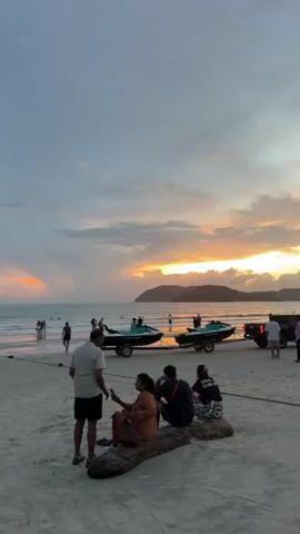 The video shows a crowded beach at sunset. In the foreground, four people are sitting on a large piece of driftwood. Two women are in the center, facing away from the camera. A man is standing to their left, facing them. Another person, seen from the back, is sitting to the right of the women.

In the middle ground, there are several people walking on the beach, some near the water's edge. Two jet skis on trailers are parked on the sand, with a few people around them. A dark-colored truck is parked further to the right.

In the background, the ocean stretches out to the horizon, with a hilly island visible in the distance. The sky is filled with dramatic clouds, illuminated by the setting sun, which casts a warm orange and yellow glow. The overall atmosphere is that of a lively evening at a tourist destination.
