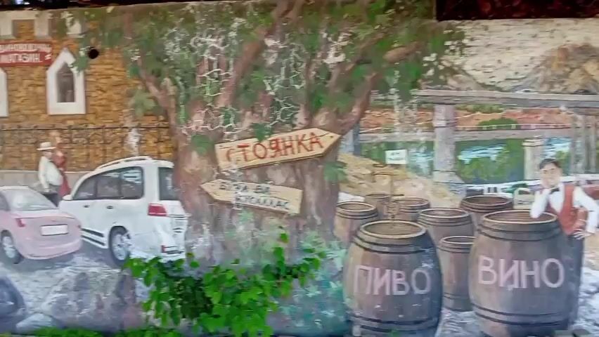 The video shows a wall with a large, detailed mural. The mural depicts a rural scene with mountains in the background. A bridge with traffic runs across the scene. There is a herd of cattle in the foreground, with a man leaning against a large wooden barrel labeled "Вино" (wine). Other barrels, labeled "Пиво" (beer), are also visible. A wooden sign with arrows points in different directions, indicating "Къща" (house) and "Янка." The mural includes trees, buildings, and cars, creating a busy, rural landscape. A white car is parked in front of the mural on the left side of the screen, partially obscured by bushes.