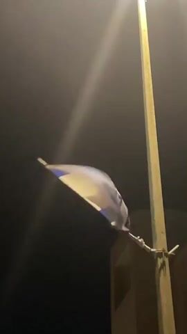 At night, a white and blue flag, likely the flag of Israel, is seen fluttering vigorously in strong wind. The flag is attached to a lamppost, which casts a bright light, illuminating the flag and the immediate area. The sky is dark, and part of a building is visible in the background. The distinct sound of strong wind blowing can be heard. The scene takes place in Mitzpe Ramon, Israel. No people or text are visible.